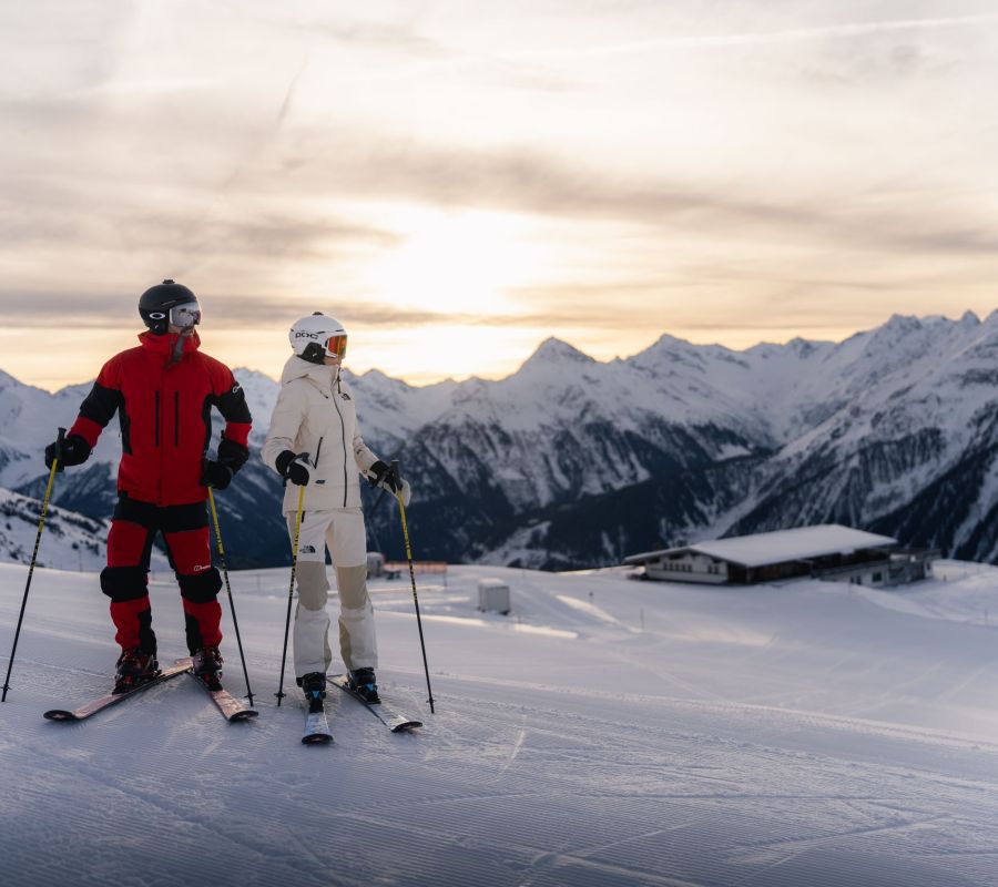 Zwei Skifahrer stehen mit ihren Skiern auf einer Piste und genießen den Sonnenuntergang in den Zillertaler Alpen.