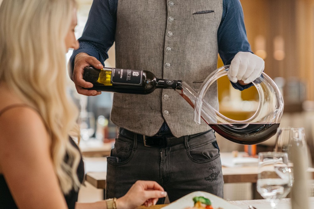 Servicemitarbeiter schenkt einer Frau am gedeckten Restauranttisch Wein ein im Alpinhotel Berghaus in Tux.