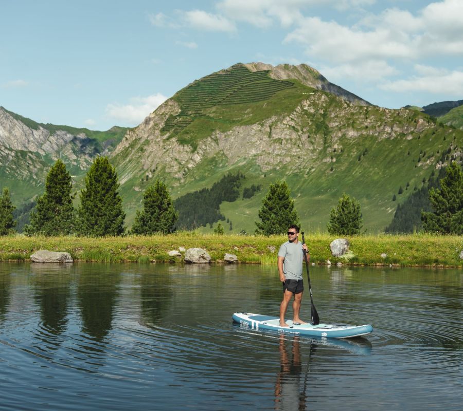 Mann beim Stand-up-Paddling auf einem Bergsee vor grünen Almen und Berggipfeln.