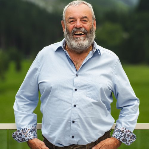 Wanderführer Sepp vom Alpinhotel Berghaus lachend im Porträt mit Lederhose vor grüner Berglandschaft.