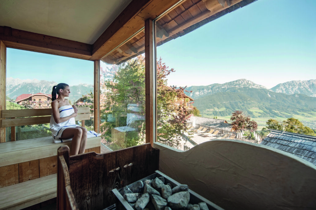 Frau sitzt in einer Sauna mit einem riesigen Panoramafenster mit Blick auf den Dachstein