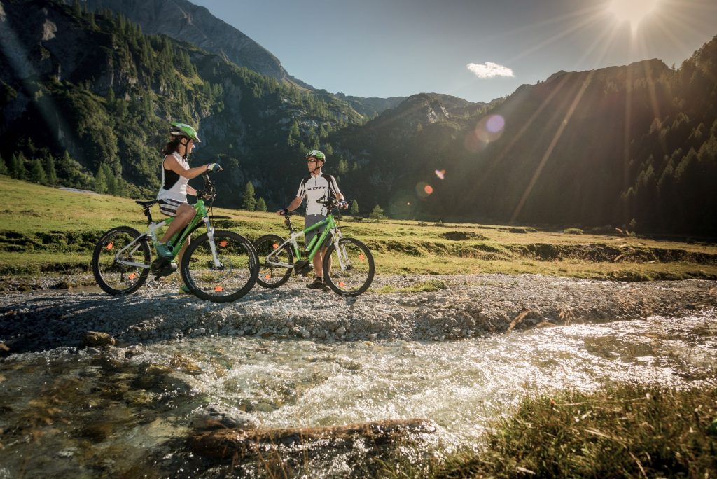 Pärchen steht mit E-Bikes im Sommer an einem rauschenden Gebirgsbach und macht Pause