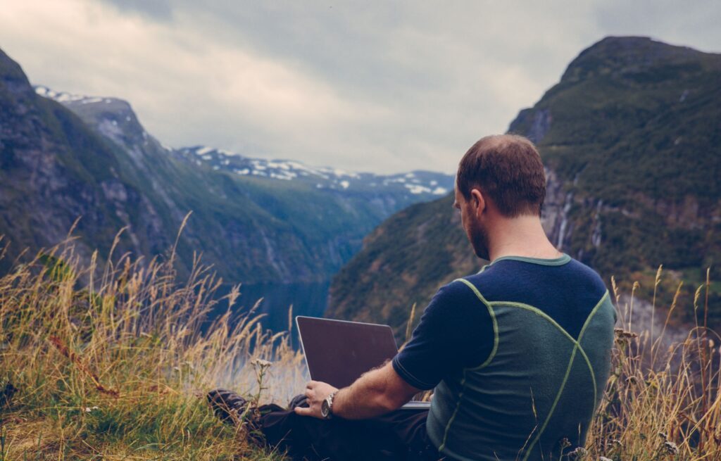 Mann sitzt mit Laptop im Gras. Panorama Berge, See im Tal
