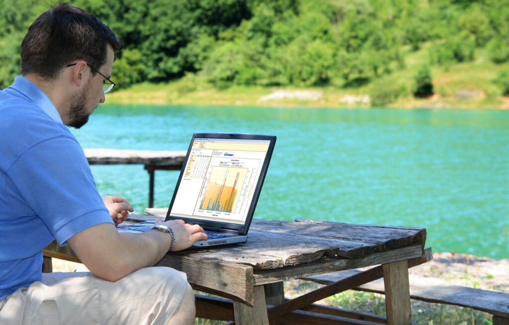 Mann sitzt an einem See und arbeitet am Laptop