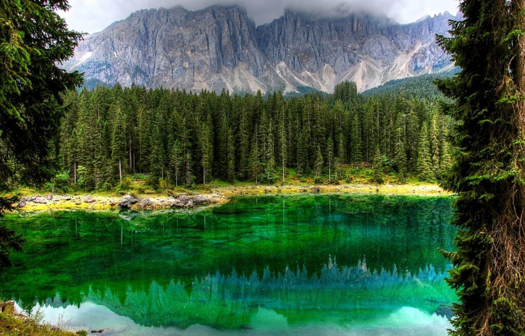 Der Karersee leuchtet grün und türkis und spiegelt den Wald und die dahinter liegenden Berge im klaren Wasser