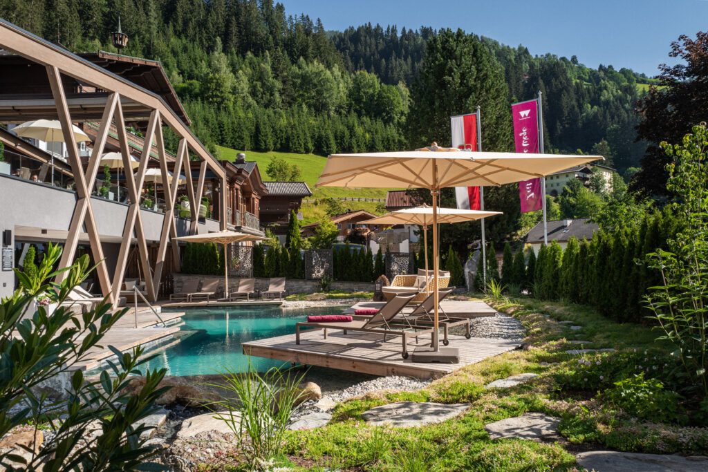 Das Natur- und Wanderhotel Gassner im Sommer mit Sonnenschirmen und türkisem Naturbadeteich