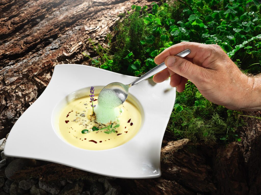 Ein eckiger Teller gefüllt mit gelber Suppe, auf die gerade ein Löffel grünen Schaums kommt