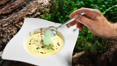 Ein eckiger Teller gefüllt mit gelber Suppe, auf die gerade ein Löffel grünen Schaums kommt© Wander- & Wellnesshotel Gassner