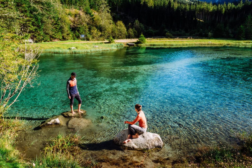 Ein Mann und eine Frau stehen beziehungsweise sitzen auf Steinen in einem türkisblauen, sehr klaren Blausee in Österreich