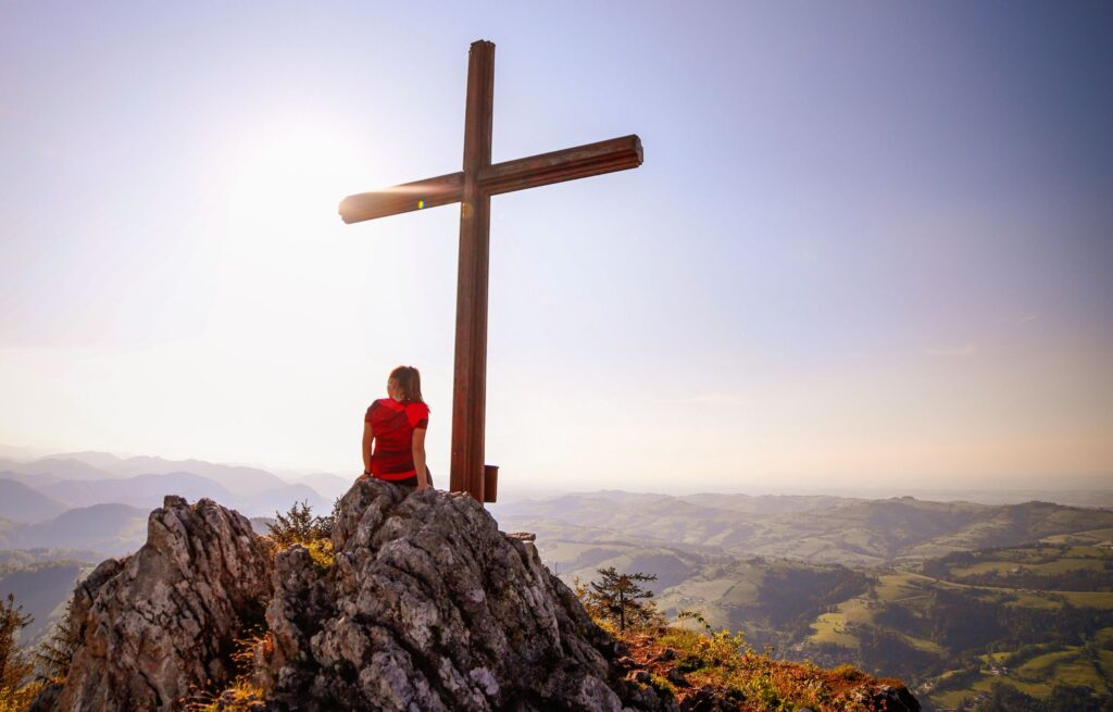 Frau sitzt auf einem Berggipfel unter einem großen Holzkreuz
