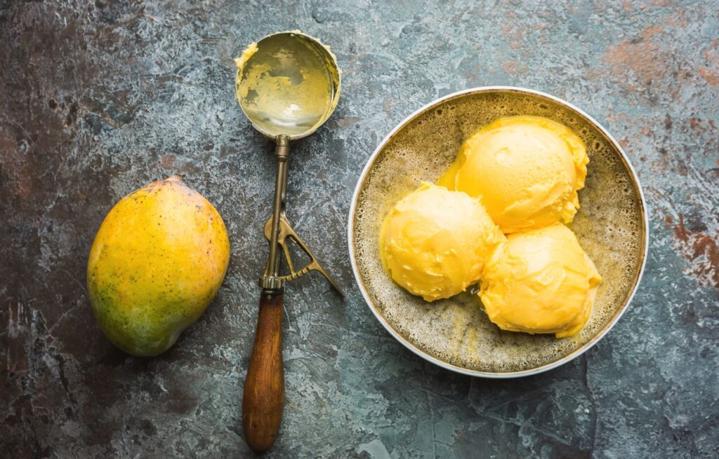 Drei Kugeln Mangoeis in einer Schüssel mit einem Eislöffel und einer Mango