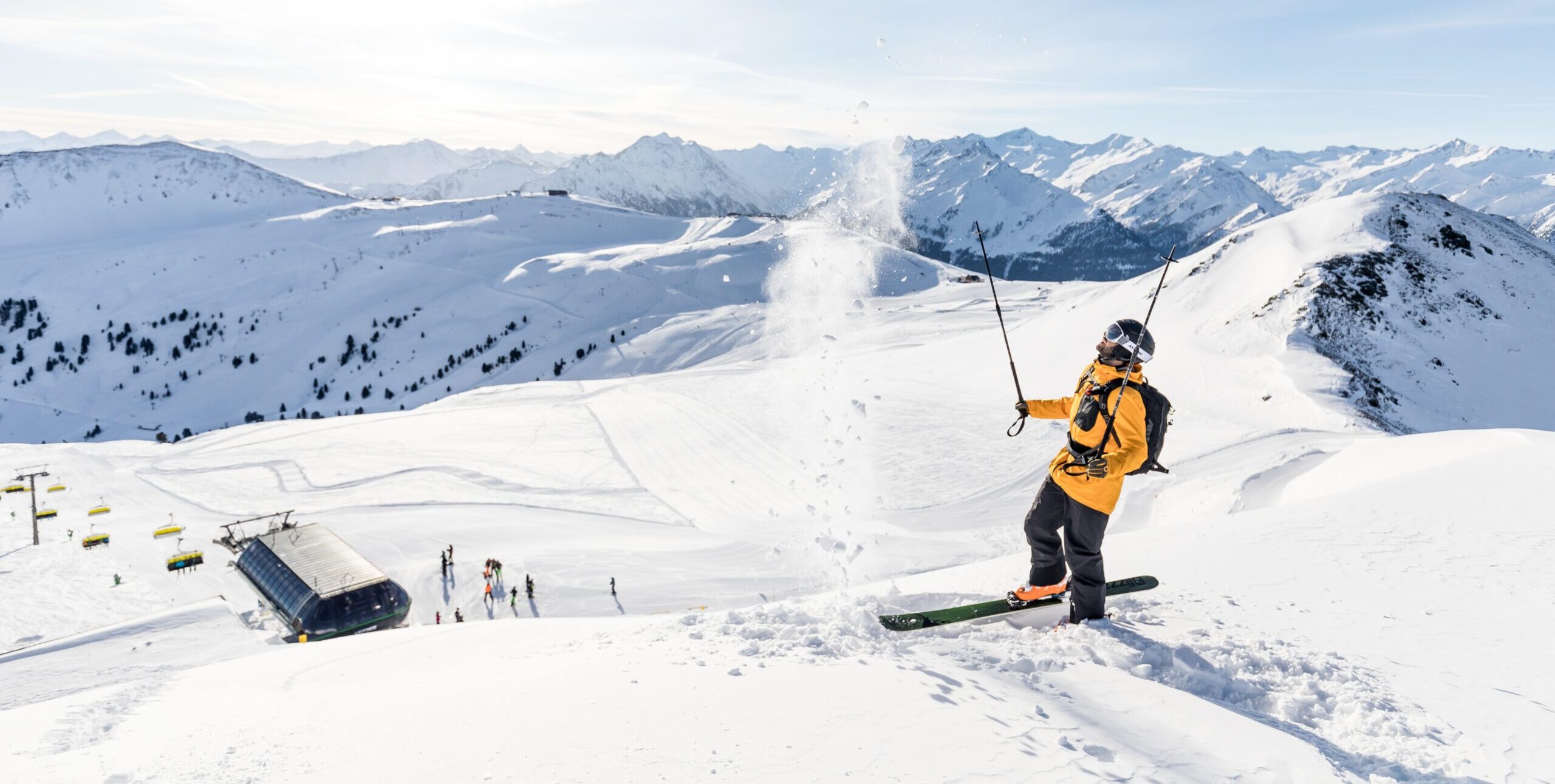 Skifahrer steht auf einem Hügel unten ist der Lift und eine verschneite Bergkulisse - er wirft den Schnee mit den Skistecken in die Luft