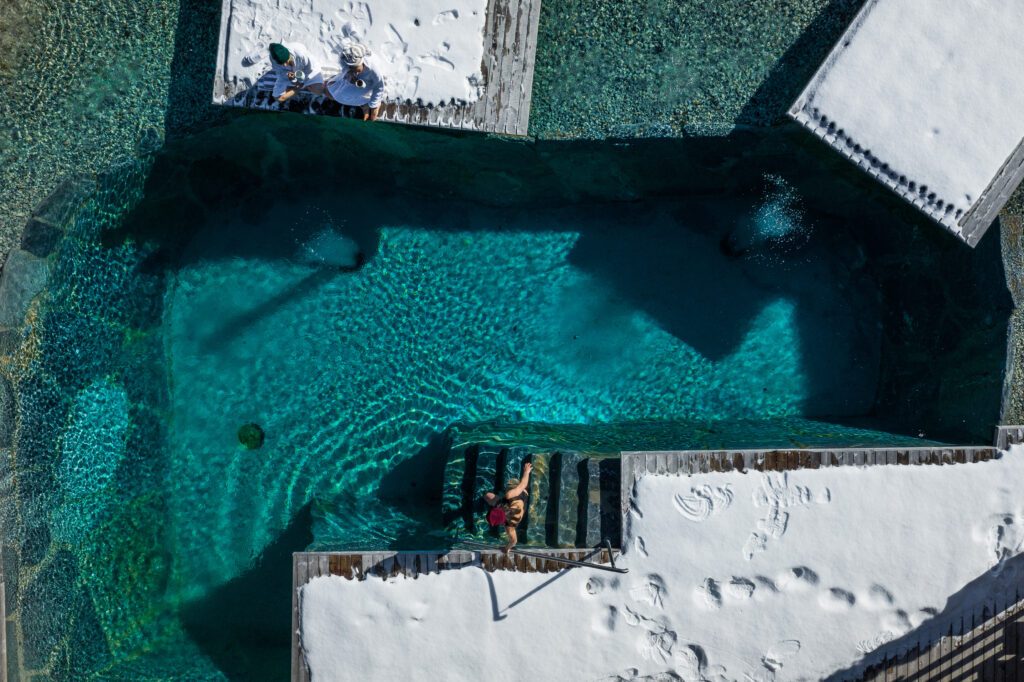 Wander- & Wellnesshotel Gassner Bild vom Pool von oben Schnee ist auf den Liegeflächen der Pool hat die Farbe Türkis