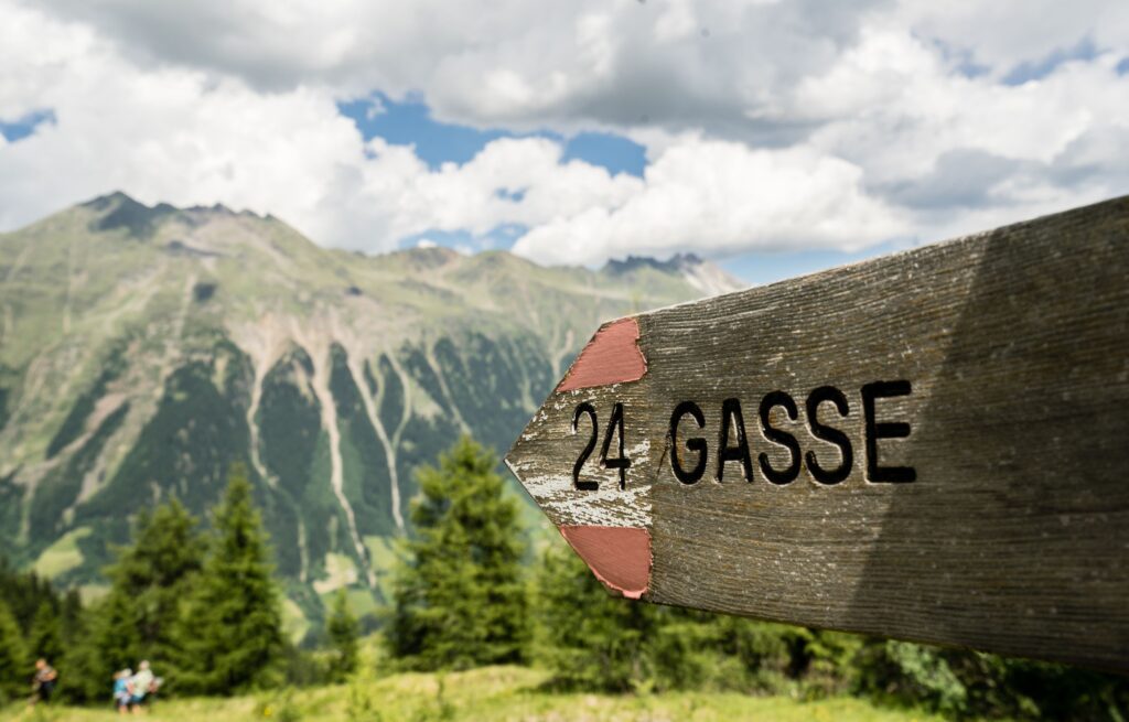 Wegweiser aus Holz mit rot-weiß-roten Streifen und der Aufschrift Gasse mit Bergen vom Südtiroler Ridnauntal im Hintergrund