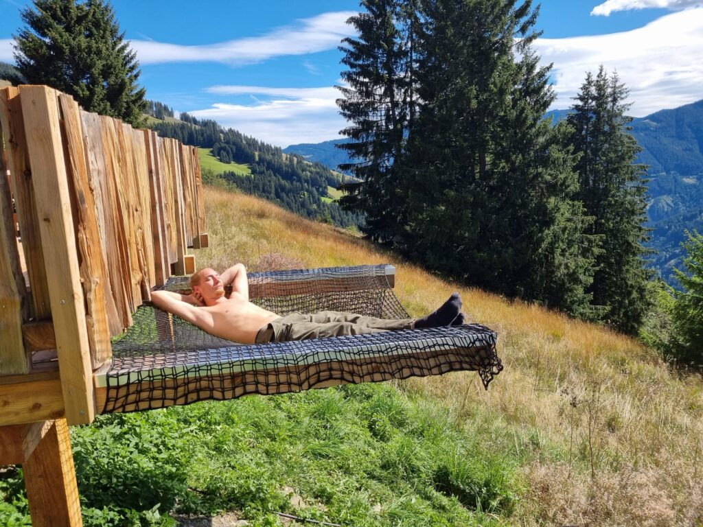Mann liegt in einem Netz am Balkon einer Almhütte im Salzburger Land und genießt.