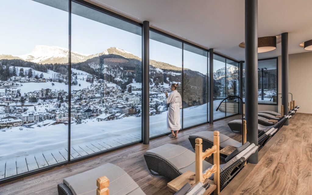 Frau im Bademantel steht am Panoramafenster des Ruheraums vom Wanderhotel Rainell Dolomits Retreat und schaut in den Winter